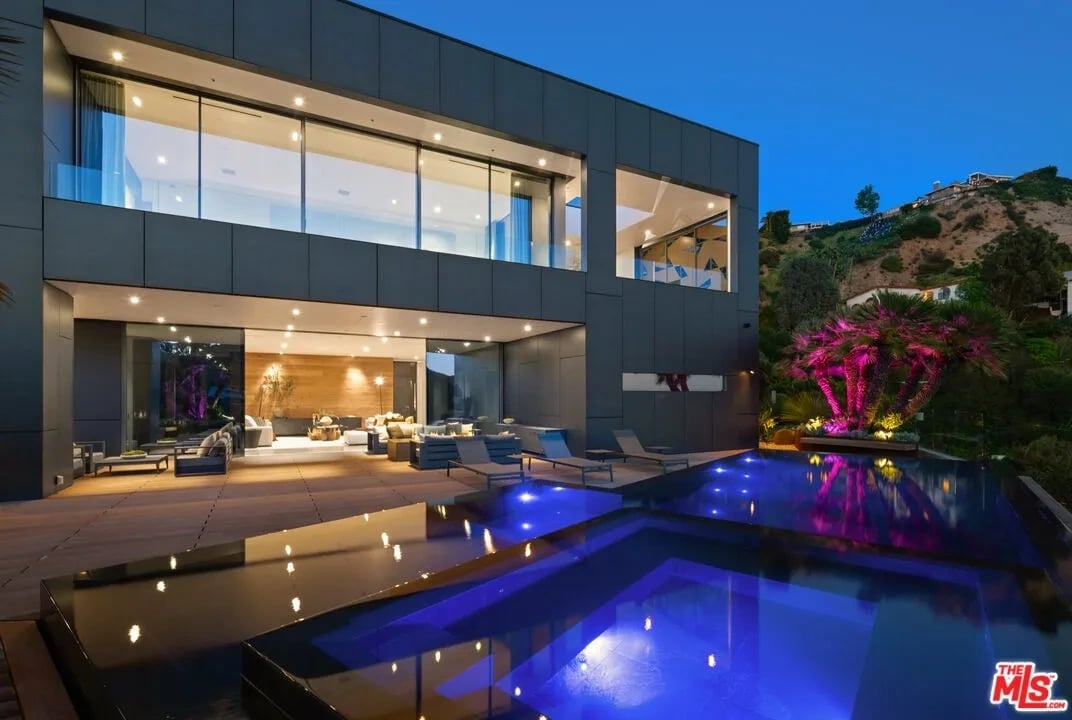 Modern glass-fronted home beside an illuminated pool and furnished patio at dusk.