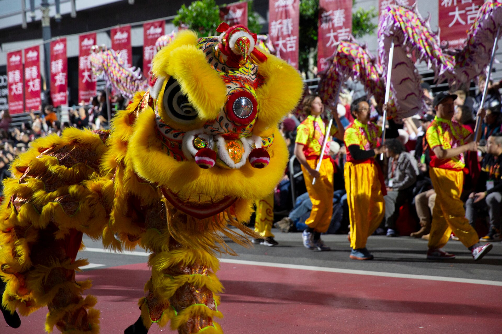 A traditional Chinese lion dance performance during a festive parade. A bright yellow lion costume with intricate patterns and fur moves along the parade route, engaging with the crowd. Performers in matching yellow and red outfits carry decorative banners and props, while onlookers line the streets, with red banners featuring Chinese characters in the background.