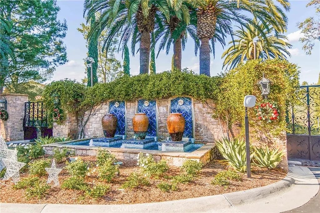 Landscaped gated entrance with a blue-tiled fountain and three urns beneath palm trees.