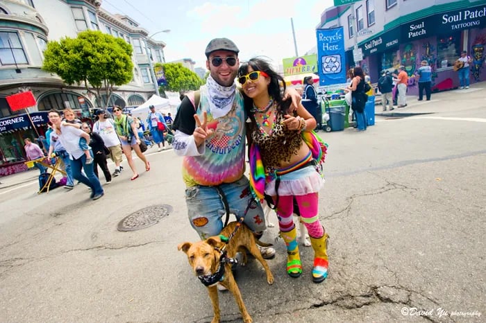 A man and woman in colorful, bohemian clothing pose with their dog on a street during the Haight-Ashbury Street Fair.