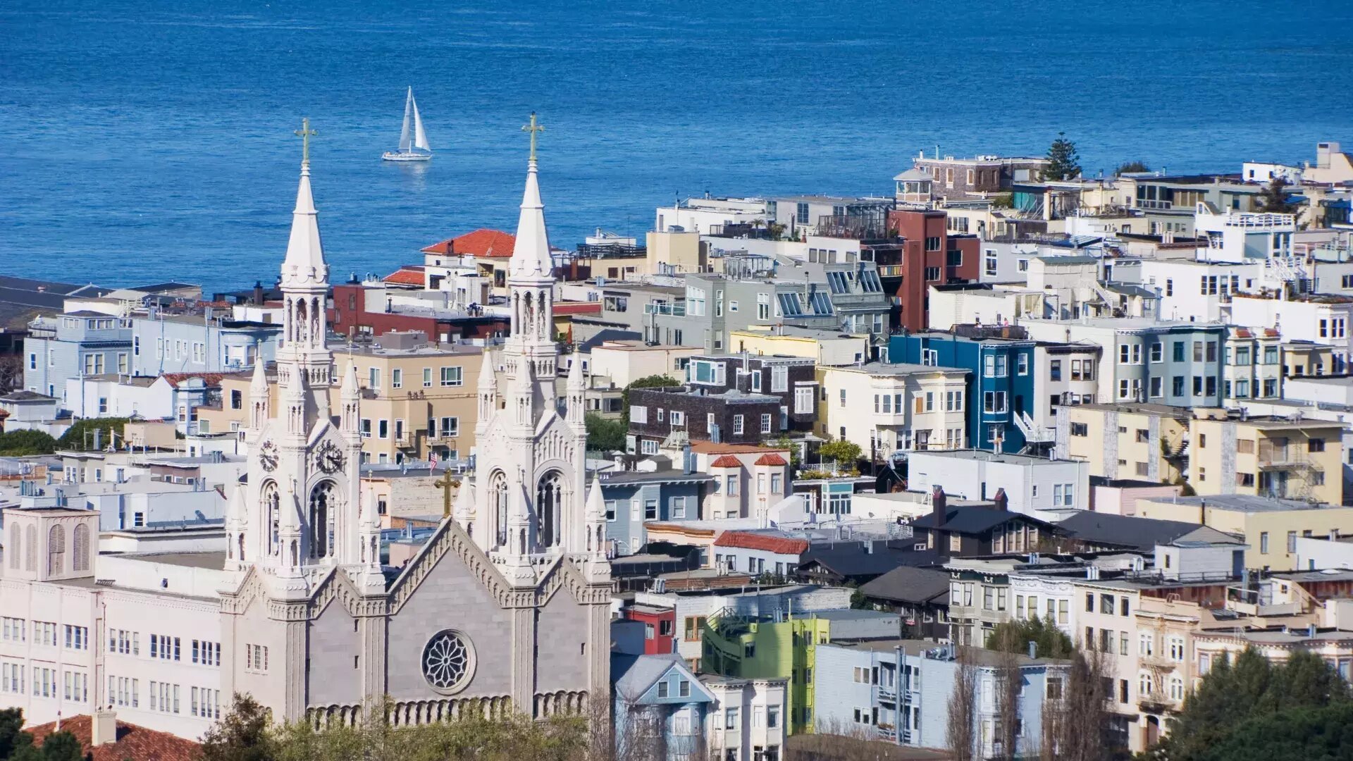 A scenic view of North Beach, San Francisco, featuring Saints Peter and Paul Church with its twin spires in the foreground. Behind the church, colorful residential buildings fill the hillside, leading to the blue waters of the San Francisco Bay, where a sailboat glides in the distance.