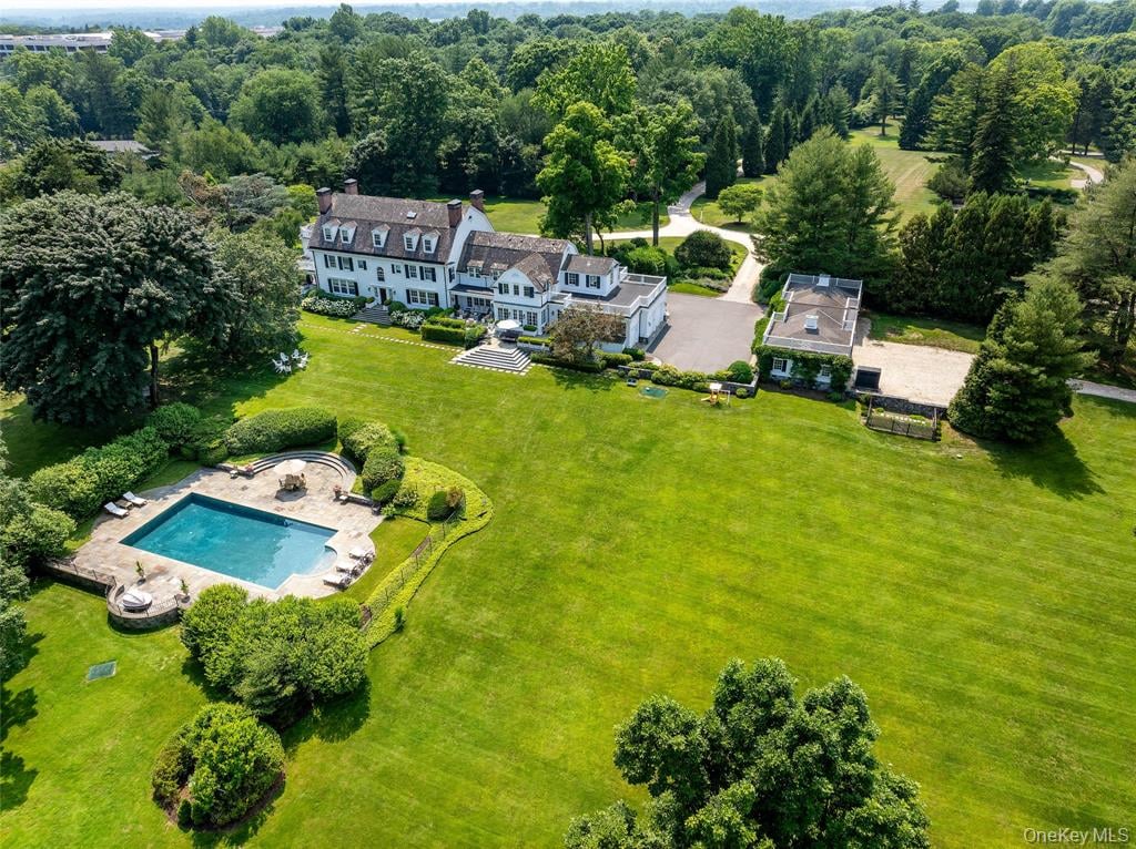 Aerial view of a white estate with a swimming pool, broad lawns, and wooded grounds.