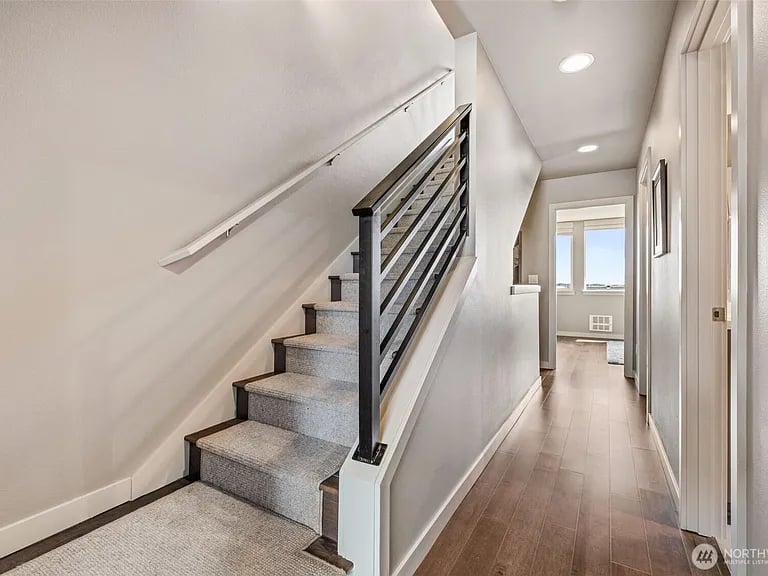 Carpeted staircase with black railing beside a bright hallway with wood-look flooring.