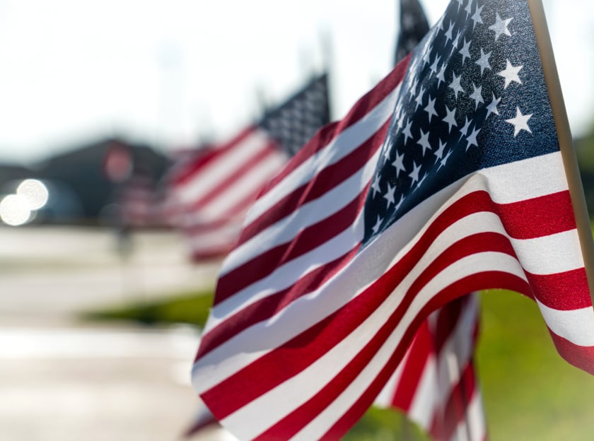 Close-up of several American flags displayed along a residential street, with the nearest flag in sharp focus and the others softly blurred in the background, creating a patriotic scene with a shallow depth of field