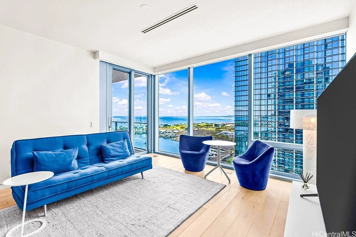 Living room with blue seating and floor-to-ceiling windows overlooking the ocean and nearby high-rises.