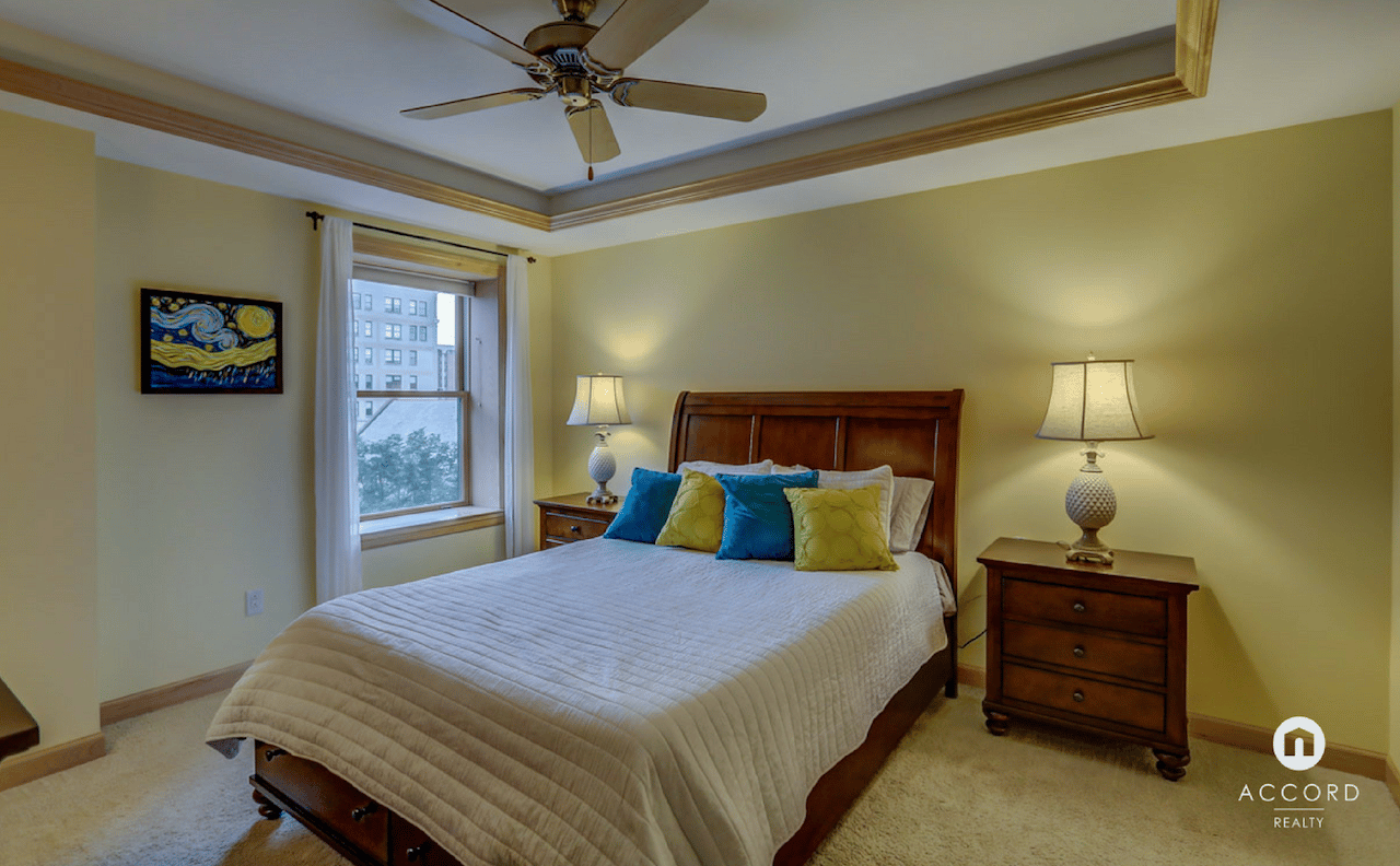 Furnished bedroom with a dark wood bed, colorful pillows, tray ceiling, and window overlooking nearby buildings.