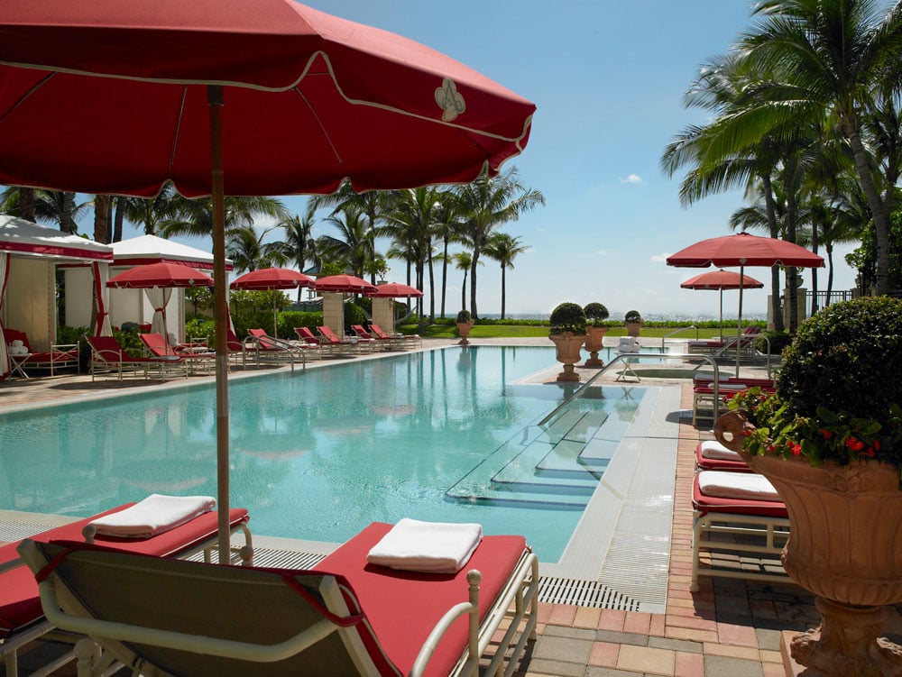 Resort swimming pool lined with red umbrellas and loungers, with palm trees and ocean beyond.