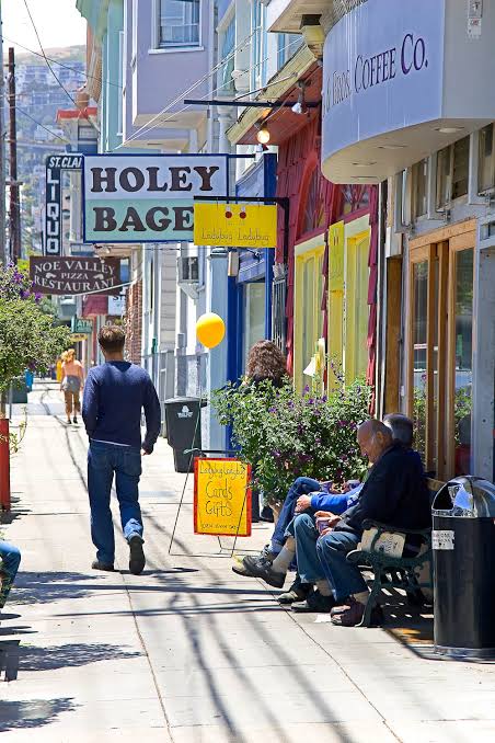 San Francisco neighborhood sidewalk with colorful storefronts, café seating, and Holey Bagel signage.