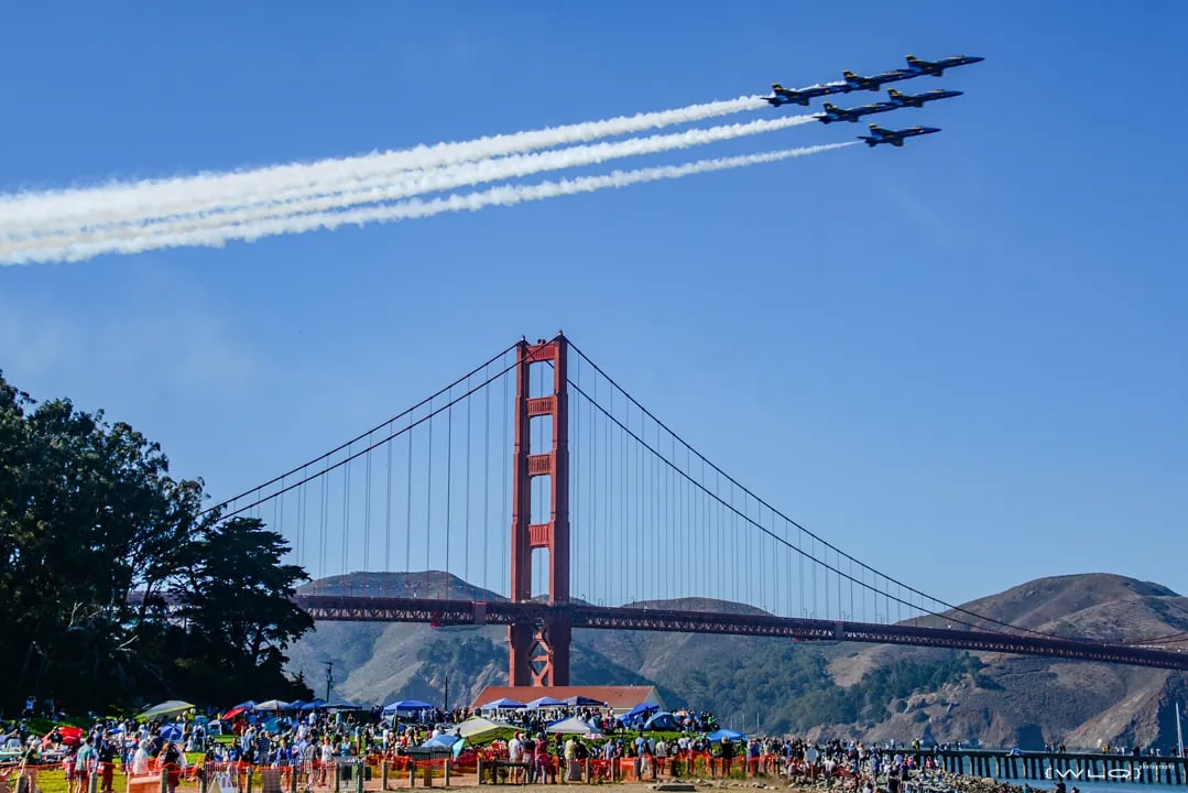 Blue Angels flying in delta formation with white contrails over Golden Gate Bridge, large crowd gathered at Crissy Field waterfront with Marin Headlands in background