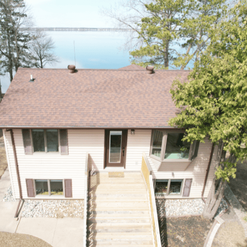 Two-story house with a broad wooden staircase, mature trees, and a lake behind it.