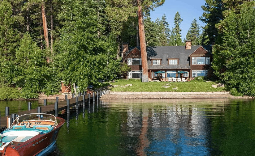 Lakefront home among tall pines, with a dock and wooden motorboat on the water.