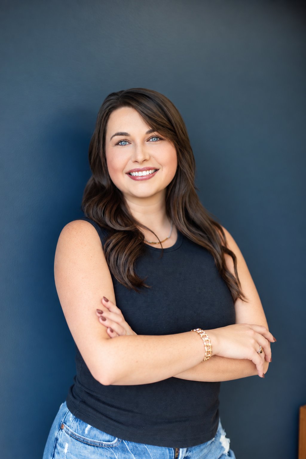 Smiling woman with long brown hair poses with arms crossed against a dark blue wall.