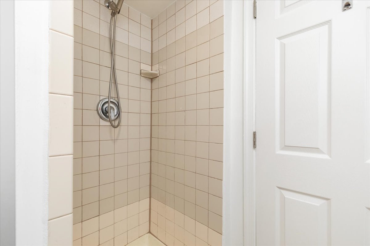 Tiled shower with a handheld showerhead, chrome controls, and a small corner shelf beside a white door.