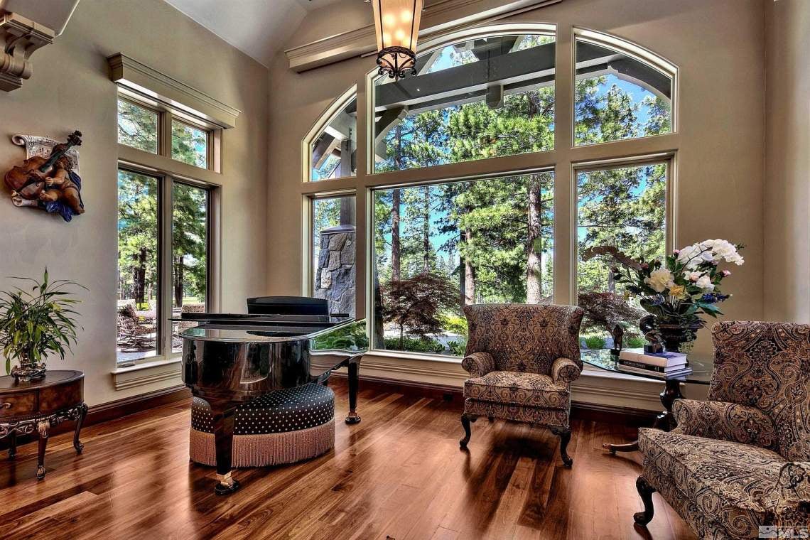 Living room with a grand piano and tall arched windows overlooking pine trees.