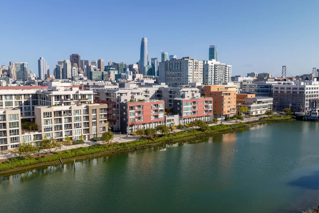 An aerial view of the Mission Bay neighborhood in San Francisco, with waterfront buildings and the downtown skyline in the background.