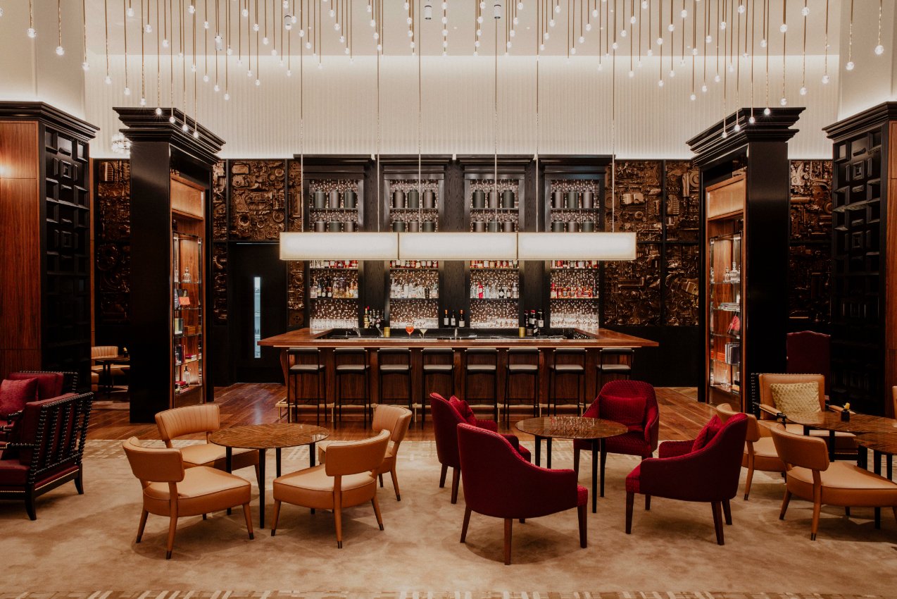 Hotel bar lounge with a long wood bar, bottle shelves, and red and tan seating.