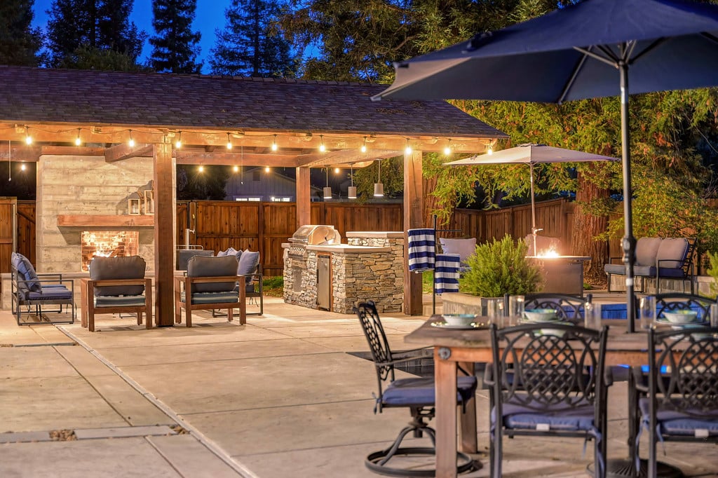 Covered outdoor kitchen and patio with stone grill, fireplace, pool, and dining seating at night.