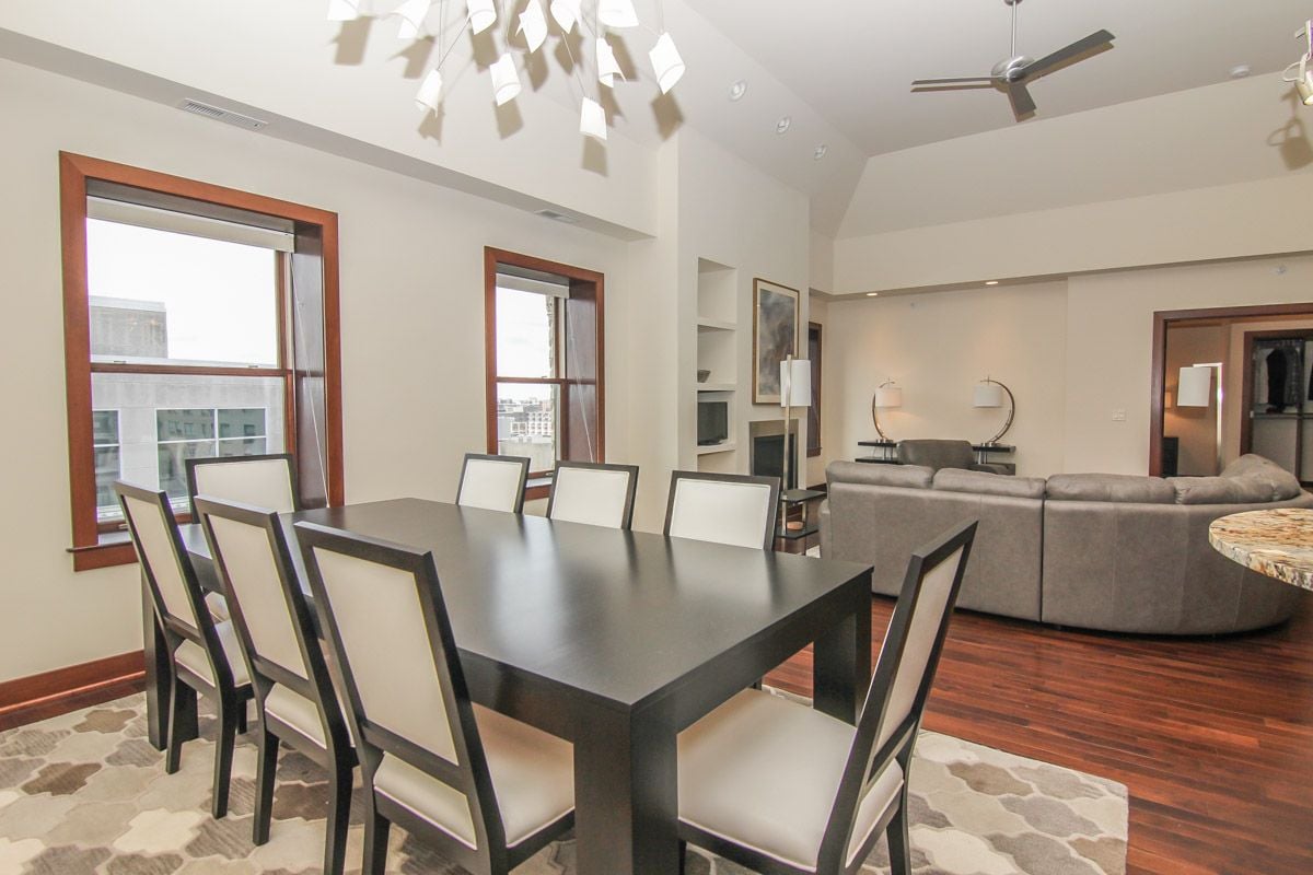Open-plan dining and living area with a dark dining table, gray sectional, and tall windows.
