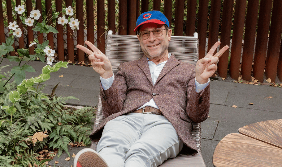 Man in glasses and a Cubs cap sitting outdoors, smiling and making peace signs.