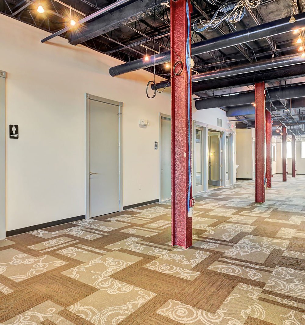Empty commercial interior with patterned carpet, exposed ceiling ducts, and red steel columns.