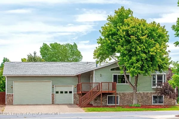 Green split-level home with a two-car garage, raised wooden porch, and large leafy tree.