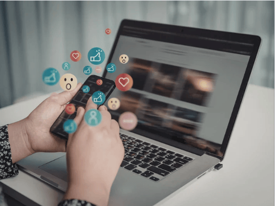 Hands using a smartphone beside a laptop, with social media reaction icons floating above.