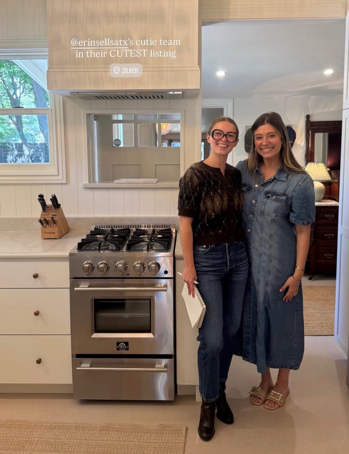Two smiling real estate team members pose beside a stainless-steel range in a bright kitchen.