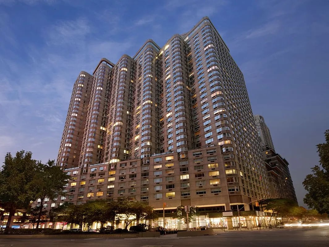 Residential condominium tower with rounded window bays and illuminated ground-floor storefronts at dusk.