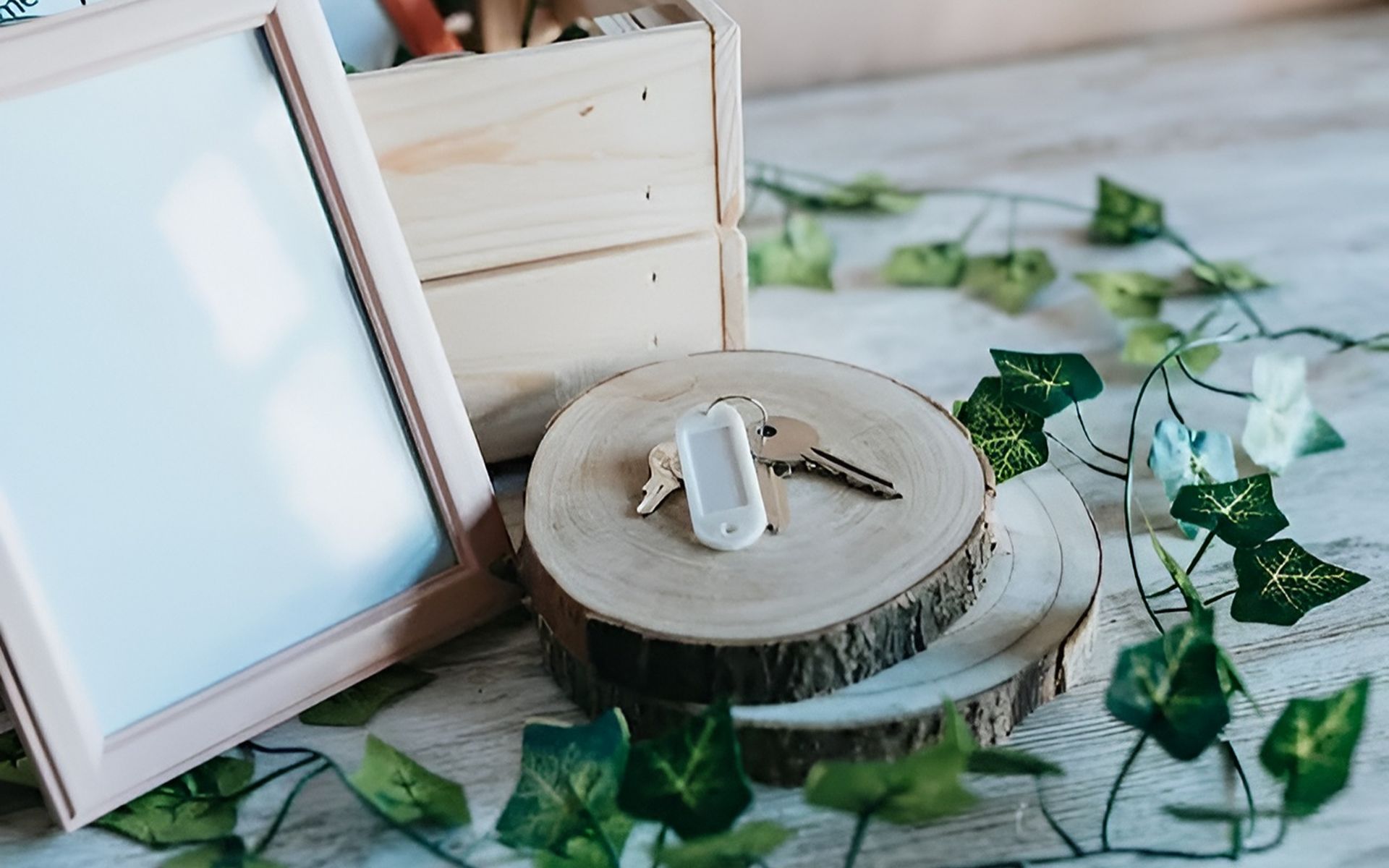 House keys and a white key fob on a wood slice beside a blank frame and ivy.