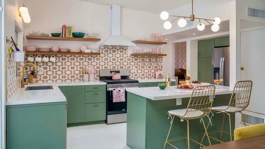 A remodeled midcentury kitchen in Palm Springs with sage green slab cabinets, a white-topped island with brass wire stools, teak open shelves, and a patterned terrazzo tile backsplash.