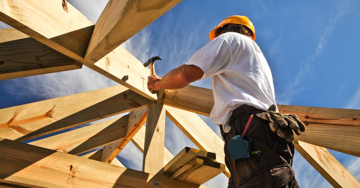 A view looking up at a worker in a hard hat with a hammer working on the roof structure of an unfinished house.