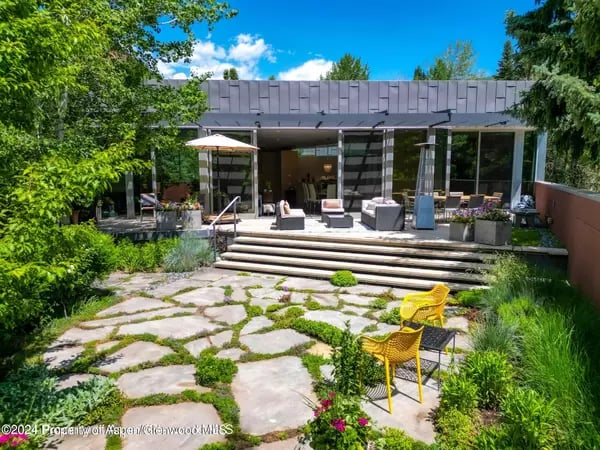 Modern home with a glass-walled patio, wide steps, and a stone garden path with yellow chairs.