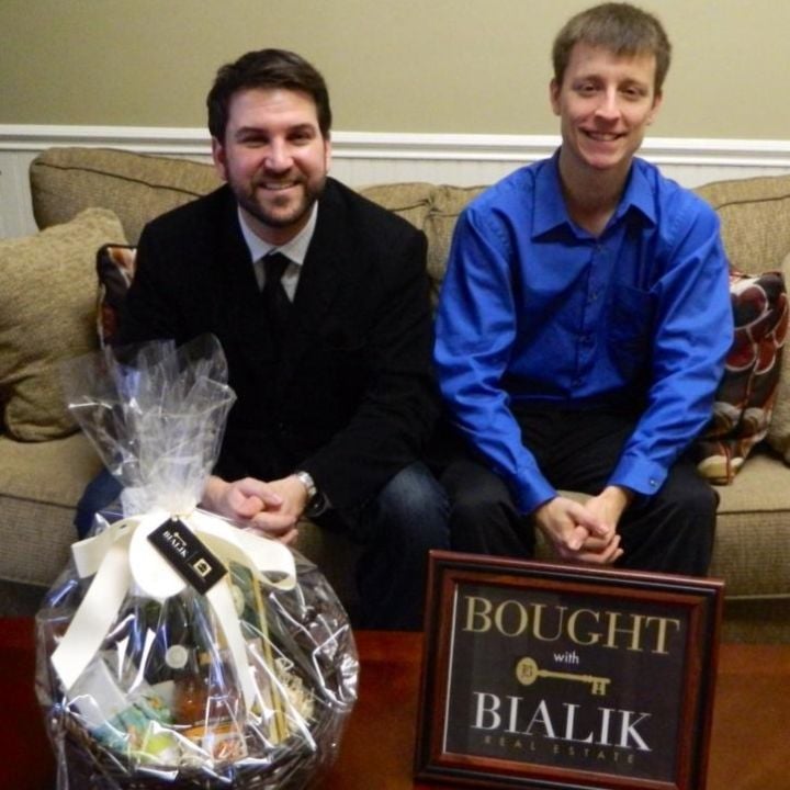 Two men celebrate a home purchase beside a gift basket and Bialik Real Estate sign.