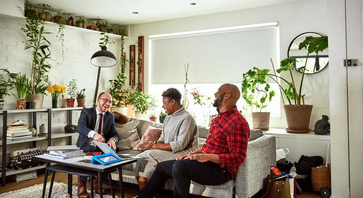 Three people sit in a cozy, plant-filled living room. A man in a suit gestures to a tablet, engaging two others. The mood is positive and collaborative.