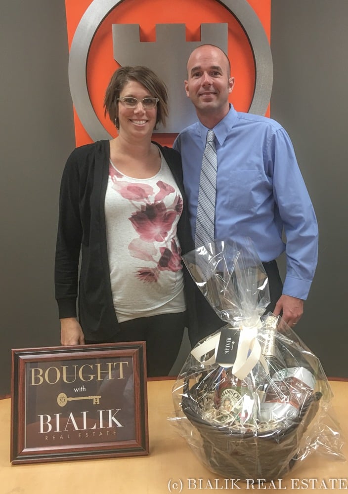 A couple poses with a gift basket beside a Bialik Real Estate home-purchase sign.