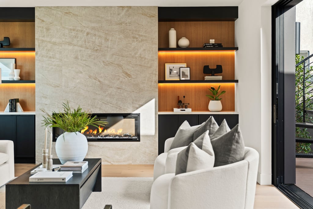 Living room with a stone fireplace, illuminated wood shelving, and white seating beside sliding glass doors.