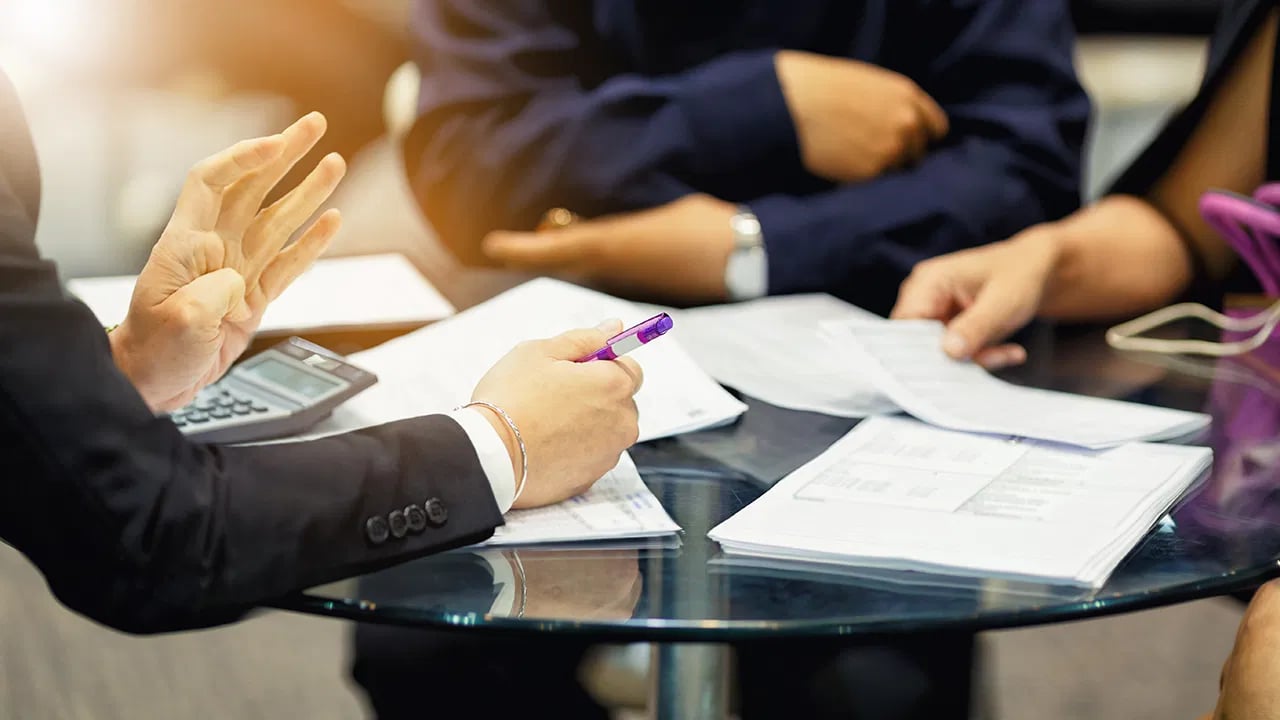 People review paperwork around a glass table as one person writes with a purple pen.