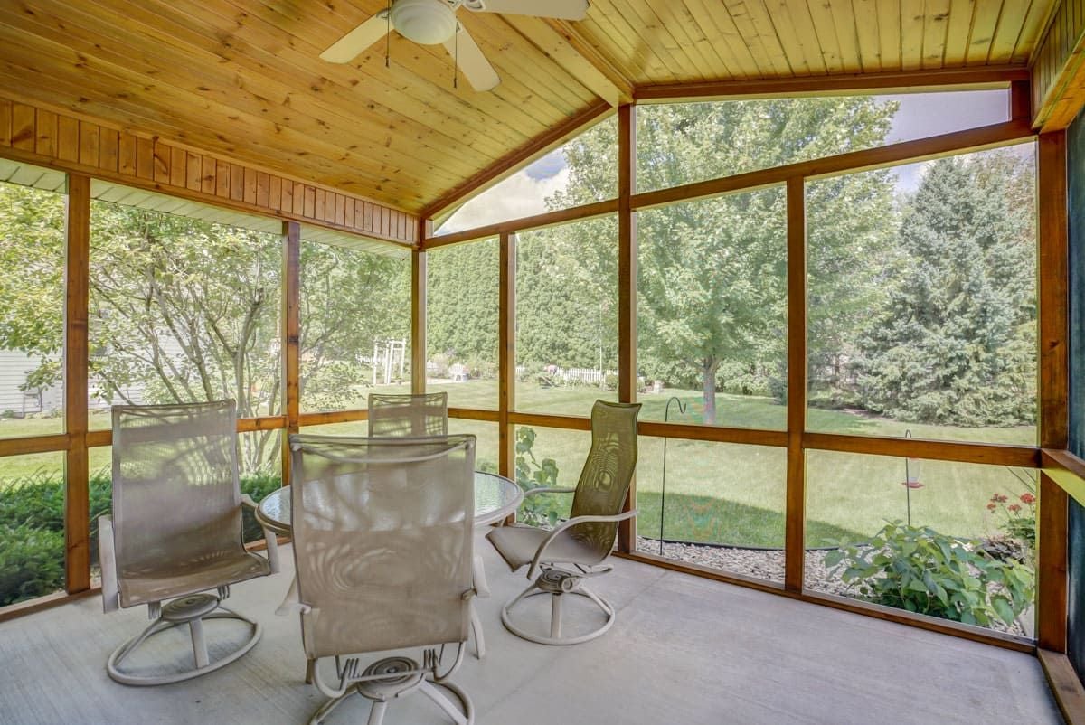 Enclosed sunroom with a knotty-pine ceiling, patio seating, and windows overlooking a leafy backyard.