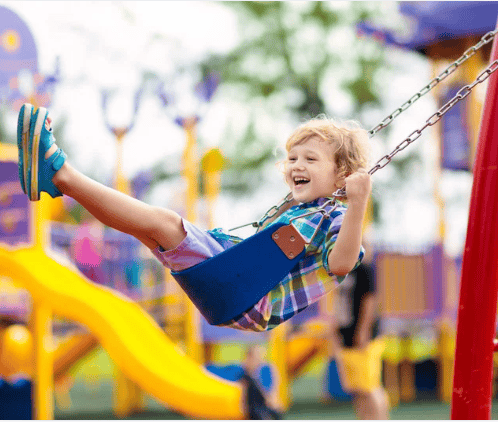 Child laughing on a playground swing beside a bright yellow slide.