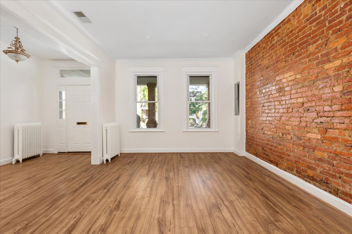 Vacant room with wood flooring, an exposed brick wall, two windows, and white radiators.