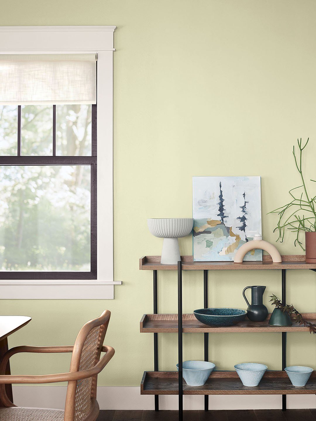 A room with walls painted Celery SW 6421, a black-framed window, and an open wood-and-metal shelf holding ceramics and plants.