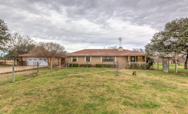 Single-story brick house with a reddish-brown roof, fenced grassy yard, and mature trees.