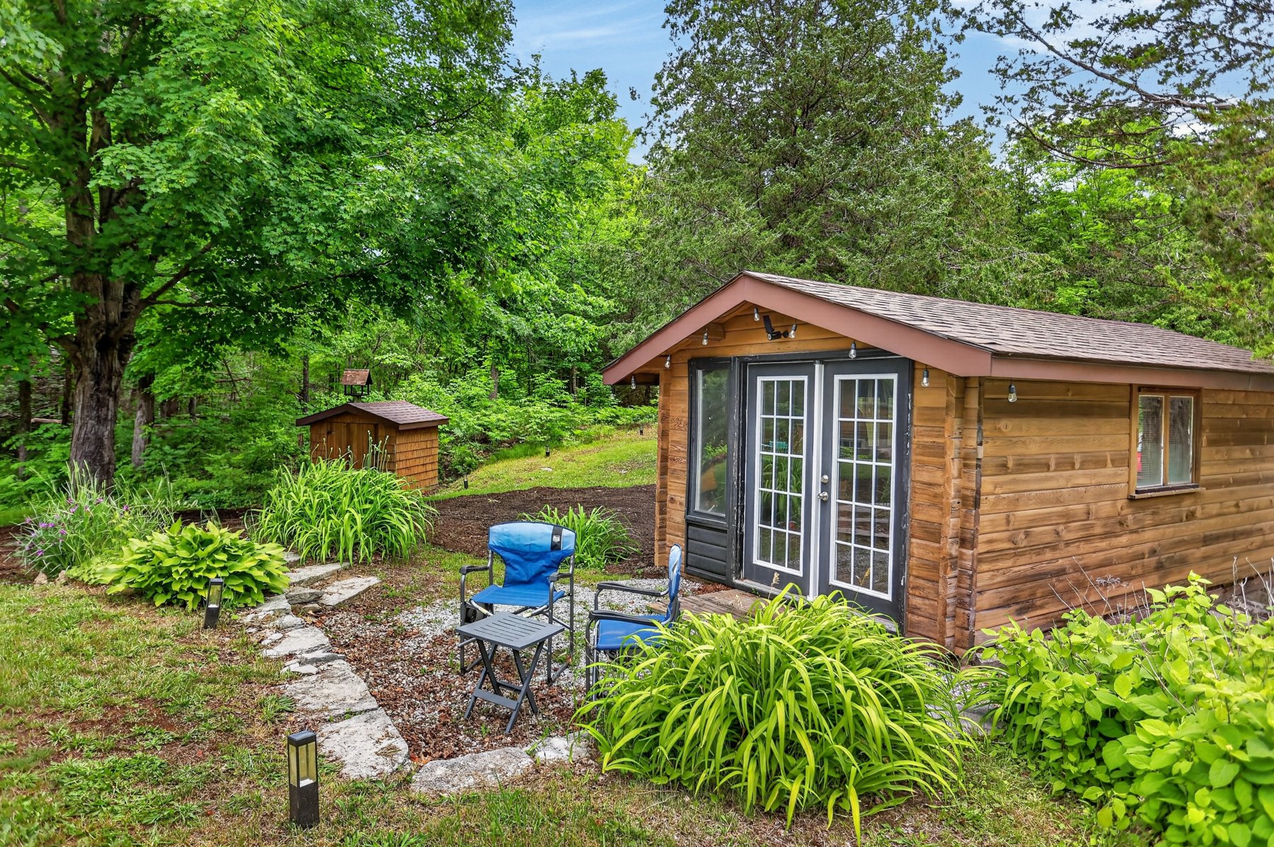 Wood-sided cabin with glass doors and blue patio chairs in a leafy garden.