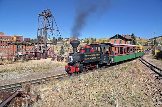 Steam locomotive pulling colorful passenger cars past a historic mining structure.