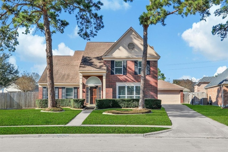 Two-story red-brick home with a gabled roof, attached garage, and mature pine trees.
