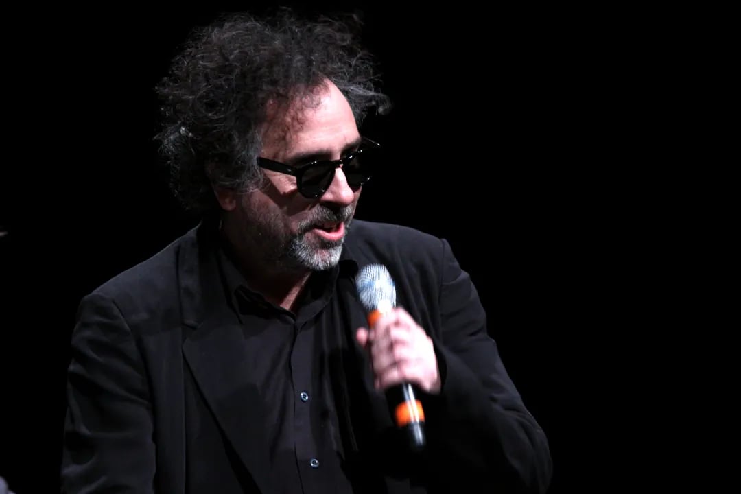 Black-and-white photo of Tim Burton on stage, holding a microphone close to his mouth, wearing dark sunglasses, a black suit jacket over a dark shirt, with his signature wild curly hair illuminated against a pitch-black background.