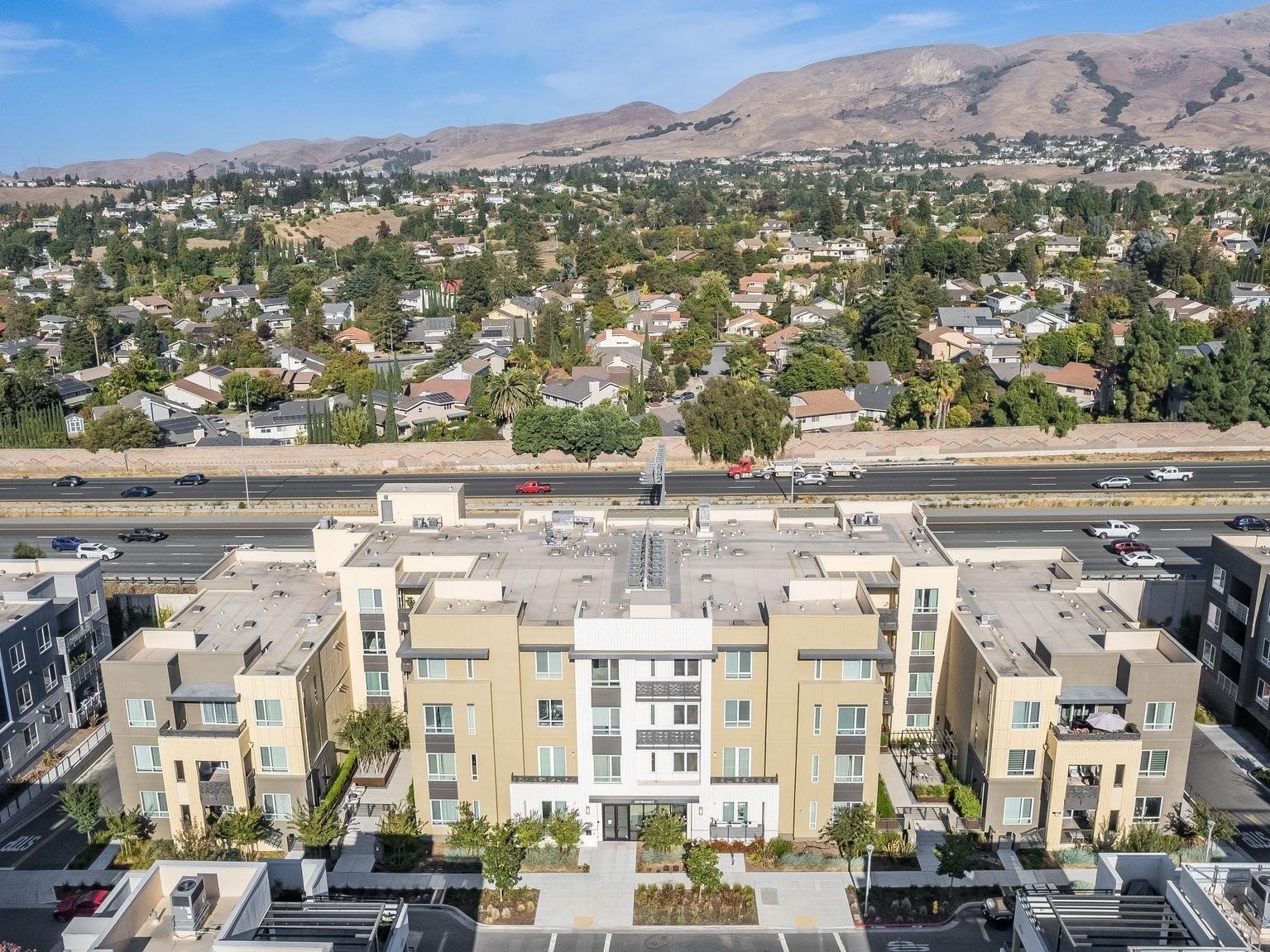 Aerial view of a modern apartment complex beside a highway, with hillside homes beyond.