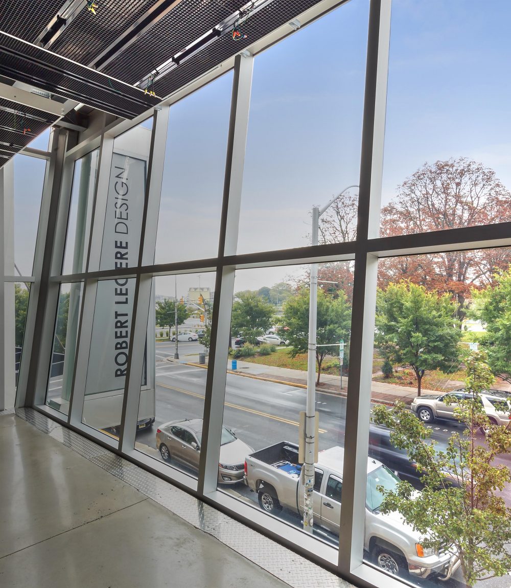 Commercial interior with floor-to-ceiling windows overlooking a tree-lined street and a Robert Legere Design sign.