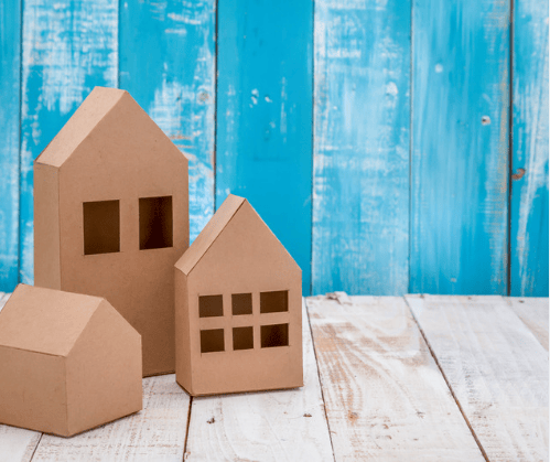 Cardboard house models with cutout windows on a white wooden surface against a bright blue backdrop.