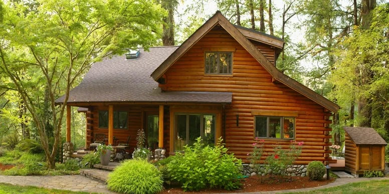 Log home with a covered porch, stone patio, and landscaped shrubs among leafy trees.